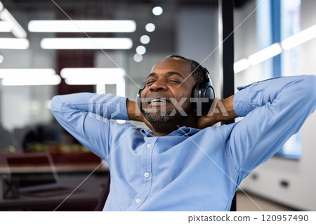 Mature African American businessman relaxing with headphones in office. Enjoying music, feeling stress-free. Calm expression suggests contentment and peace during work break. Perfect for relaxation. 120957490