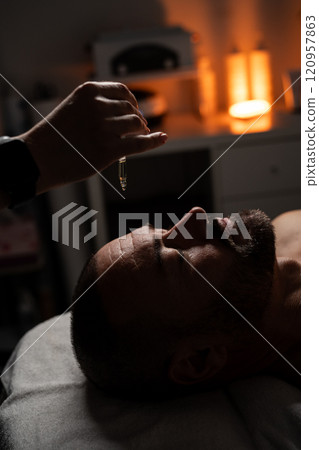Oil is dripped onto a man's forehead for massage. Close up of a handsome man enjoying facial massage at spa center. Oil is dripped onto a man's forehead for massage. Close up of a handsome man enjoying facial massage at spa center. 120957863