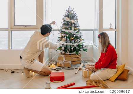 Husband and wife couple lover decorating Christmas tree together while man setting up the tree and woman packing the box. 120959617