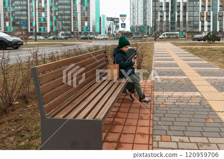 Young boy sitting on a park bench with a smartphone in a modern urban area during a cloudy day 120959706
