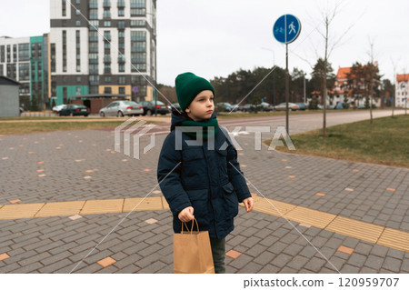 Young boy wearing green accessories stands at a crosswalk in an urban area during a cloudy day 120959707
