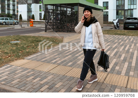 Young woman walks on a city sidewalk while talking on a phone and carrying a bag during a cool day Young woman walks on a city sidewalk while talking on a phone and carrying a bag during a cool day 120959708