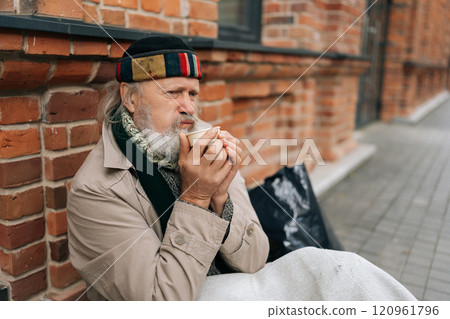Portrait of tired retired man sitting outside on city street, sipping warm drink from paper cup, evoking feelings of compassion and empathy. Frozen aged tramp male looking sad and lost. 120961796