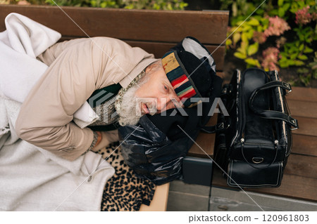 High-angle view of tired sad aged homeless man lying on bench outside, seeking refuge from elements. Sad beggar aged male bundled up in warm clothing lying with bag of belongings. 120961803