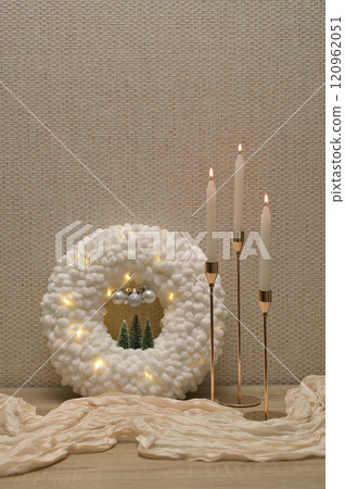 Christmas decor, white soft plush wreath with lights, three lit candles in golden holder, beige cloth on wooden table 120962051
