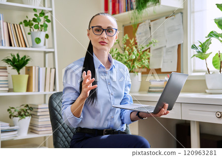 Middle aged business serious woman with laptop in hands looking at camera, in home office Middle aged business serious woman with laptop in hands looking at camera, in home office 120963281