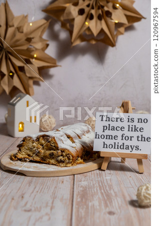 Note with saying THERE IS NO PLACE LIKE HOME FOR THE HOLIDAYS.Traditional German Christmas stollen cake in background of handmade festive decor. Winter holiday treats. Vegan baking recipe 120967594