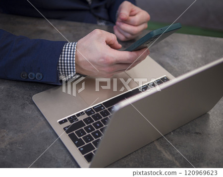 Focused businessman in modern office environment, using smartphone and laptop while dressed in blue suit and checkered shirt 120969623