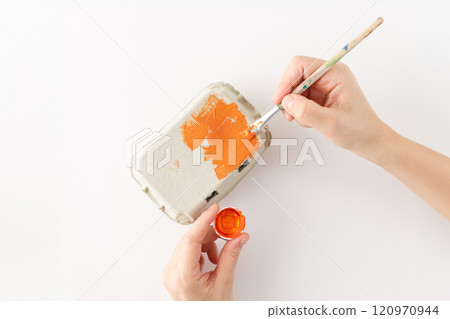 step in creative process. Painting an egg carton orange with a brush for a DIY craft project on a white background. Hands crafting in progress. Creative recycling and small kids activity concept, 120970944
