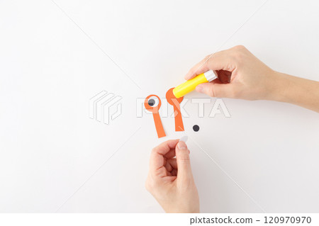 top down view of hands glue eyes details, a yellow glue stick and orange paper cut out. part of a craft project, creating a character or object with eyes. a white circle and a black circle 120970970