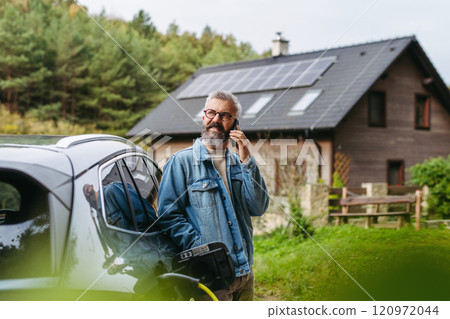 Man charging electric car in front of his house while making phone call. House with solar panel system on roof behind him. Man charging electric car in front of his house while making phone call. House with solar panel system on roof behind him. 120972044