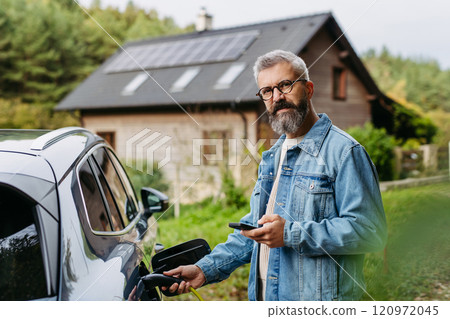 Man charging electric car in front of his house, plugging the charger into the charging port. House with solar panel system on roof behind him. 120972045