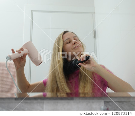 A woman is expertly styling her hair using a blow dryer and brush while in the bathroom A woman is expertly styling her hair using a blow dryer and brush while in the bathroom 120973114
