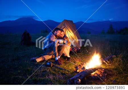 Couple hikers enjoys cozy moment by campfire, sipping hot drinks near glowing tent. Twilight sky and mountain backdrop create serene and romantic atmosphere in picturesque wilderness setting. 120973891