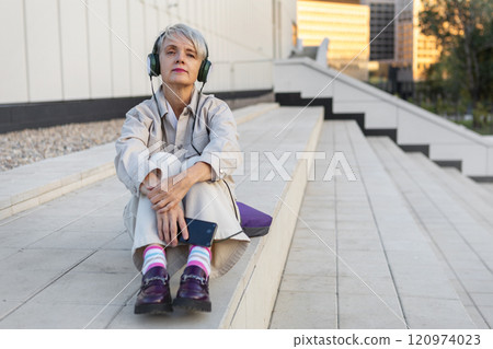 Thoughtful Lonely Trendy Mature Woman Listening To Music On Headphones While Sitting On Steps Of White Modern Building In City 120974023