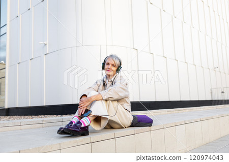 Smiling Trendy Mature Woman Listening To Music On Headphones While Sitting On Steps Of White Modern Building In City Smiling Trendy Mature Woman Listening To Music On Headphones While Sitting On Steps Of White Modern Building In City 120974043