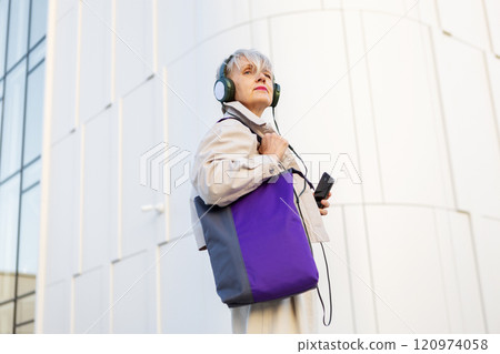 Thoughtful Older Woman with grey hair Standing Outdoors in City, Wearing Headphones, beige jacket and Holding phone, Looking Distance and Reflecting on Life on Modern Urban White building background Thoughtful Older Woman with grey hair Standing Outdoors in City, Wearing Headphones, beige jacket and Holding phone, Looking Distance and Reflecting on Life on Modern Urban White building background 120974058