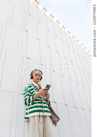 Stylish Mature Woman In Green And White Striped Sweater, Beige Pants And Headphones Listening To Music While Looking Up And Holding Phone Against White Building Wall 120974063