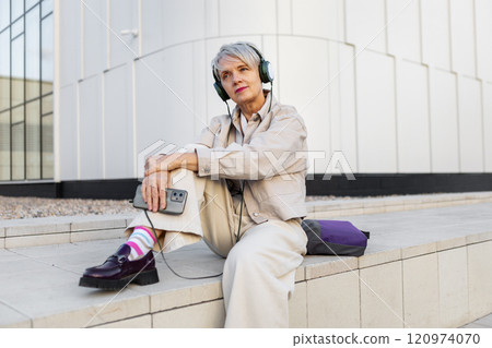 Beautiful Trendy Senior Woman with Short Hair and Headphones in Beige Clothing Sitting Outdoors in City, Listening to Music, Podcast or Audiobook Against Modern White Building 120974070