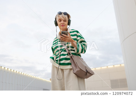 Stylish Mature Woman In Green And White Striped Sweater, Beige Pants And Headphones Listening To Music While Looking Up And Holding Phone Against White Building And Sky 120974072