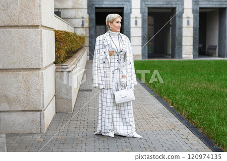 Fashionable Mature Woman with Short Hair Standing Outside in City Dressed in Stylish Checkered White Suit with Blazer and Trousers, Accessories and Handbag Showcasing Trendy Clothing and Street Style 120974135