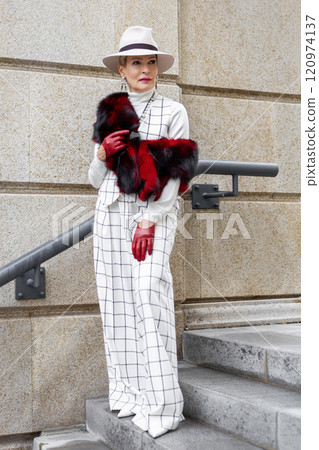 Fashionable Mature Woman Walking Outside in City Dressed in Stylish Checkered White Suit and Trousers with Fedora Hat, Faux fur Accessories Showcasing Trendy Clothing and Street Style Fashionable Mature Woman Walking Outside in City Dressed in Stylish Checkered White Suit and Trousers with Fedora Hat, Faux fur Accessories Showcasing Trendy Clothing and Street Style 120974137
