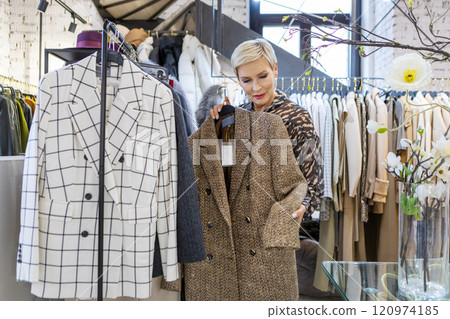 Beautiful mature woman in fashionable tweed blazer Shopping and Trying On clothes, stands near rack in luxurious boutique and smiling, confidence, style and elegance Beautiful mature woman in fashionable tweed blazer Shopping and Trying On clothes, stands near rack in luxurious boutique and smiling, confidence, style and elegance 120974185