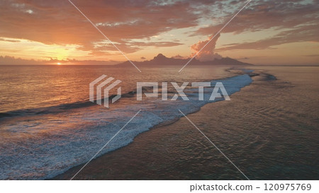 Aerial view of bright orange sunset over Moorea island in French Polynesia. Waves gently rolling the shore. Creating a picturesque and tranquil scene with warm colors reflecting on the ocean surface 120975769