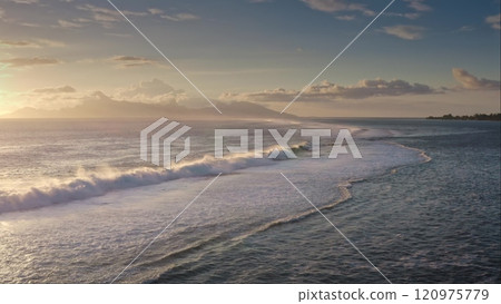 Ocean waves crashing the shore during golden sunset, with distant tropical island mountains visible on the horizon. Clouds adding depth to colorful sky, dramatic cloudscape. Nature travel background 120975779
