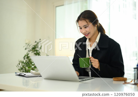 Smiling young businesswoman holding a coffee mug and working on her laptop in office Smiling young businesswoman holding a coffee mug and working on her laptop in office 120976050