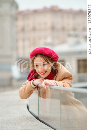 A young woman in a red beret smiles while leaning on a railing in an urban setting during daytime A young woman in a red beret smiles while leaning on a railing in an urban setting during daytime 120976243
