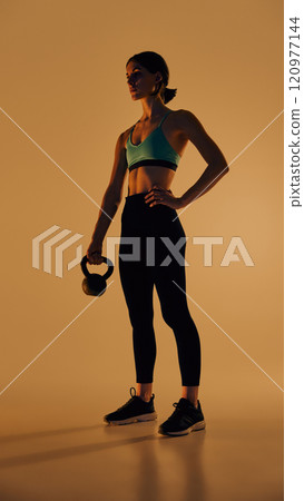 Full-length low angle view image of young slim woman in sportswear standing with kettlebell, posing against yellow background in neon light Full-length low angle view image of young slim woman in sportswear standing with kettlebell, posing against yellow background in neon light 120977144