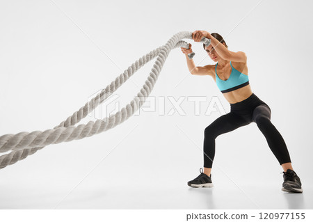 Powerful and fit young woman with slim body, in sportswear, training with battle rope against white studio background 120977155