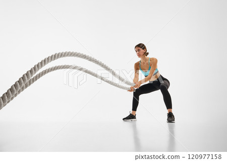 Young sportive woman with fit, muscular body training with battle rope, showing power and endurance against white studio background 120977158