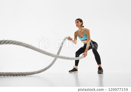 Young sportive woman with fit, muscular body training with battle rope, showing power and endurance against white studio background 120977159
