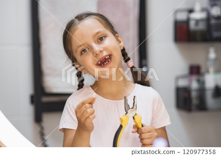 Caucasian girl smiling in bathroom holding fallen baby tooth and pliers, reflecting excitement and achievement after tooth loss. 120977358