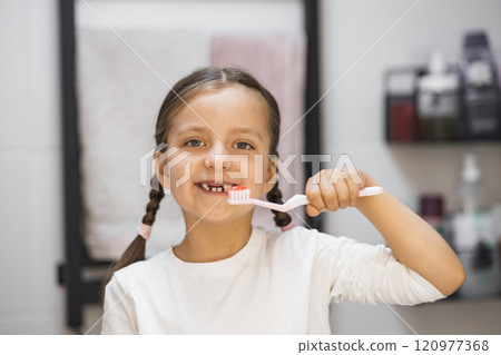 Caucasian girl with braided hair brushing her teeth and smiling at mirror in bathroom setting. Displaying missing milk teeth and engaging in daily dental hygiene routine. 120977368