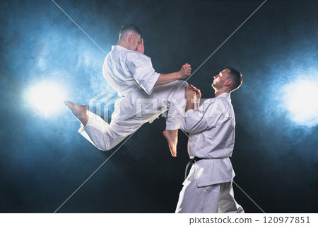 Martial arts masters on dark background. Strong man fighters in white kimono for karate martial arts fighting training in black gym. Karate fighters training punches kicks practicing fight sparring Martial arts masters on dark background. Strong man fighters in white kimono for karate martial arts fighting training in black gym. Karate fighters training punches kicks practicing fight sparring 120977851