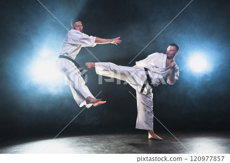 Martial arts masters on dark background. Strong man fighters in white kimono for karate martial arts fighting training in black gym. Karate fighters training punches kicks practicing fight sparring Martial arts masters on dark background. Strong man fighters in white kimono for karate martial arts fighting training in black gym. Karate fighters training punches kicks practicing fight sparring 120977857