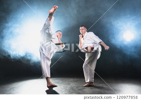 Martial arts masters on dark background. Strong man fighters in white kimono for karate martial arts fighting training in black gym. Karate fighters training punches kicks practicing fight sparring Martial arts masters on dark background. Strong man fighters in white kimono for karate martial arts fighting training in black gym. Karate fighters training punches kicks practicing fight sparring 120977858