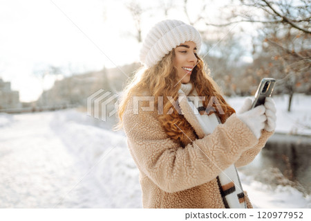 A young woman in a snowy park with a phone in her hands communicates via video and takes a selfie. A young woman in a snowy park with a phone in her hands communicates via video and takes a selfie. 120977952