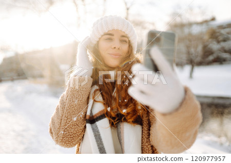 A young woman in a snowy park with a phone in her hands communicates via video and takes a selfie. A young woman in a snowy park with a phone in her hands communicates via video and takes a selfie. 120977957