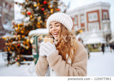 A young woman drinks a hot drink at a Christmas market decorated with festive lights in the evening. 120977961