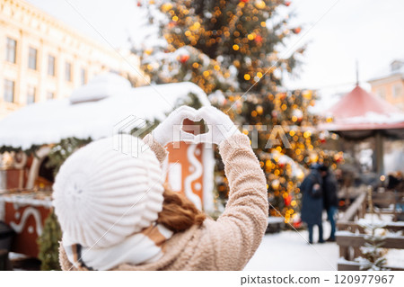 Young woman holding hands in the shape of a heart against the background of a Christmas tree. 120977967