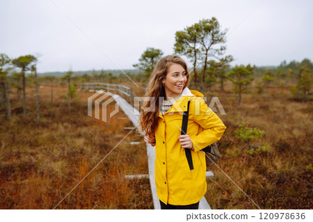 Smiling female traveler in yellow coat walks along wooden path among wild nature. Travel, vacation. Smiling female traveler in yellow coat walks along wooden path among wild nature. Travel, vacation. 120978636