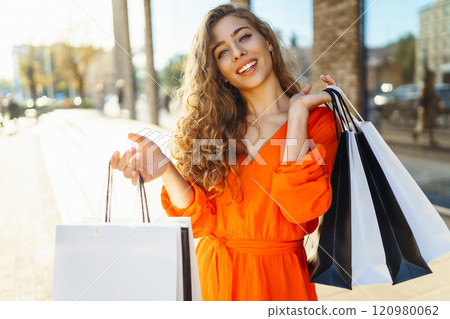 Cheerful young woman with shopping bags walking down city street. Purchases, black friday, discounts 120980062
