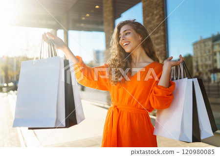 Cheerful young woman with shopping bags walking down city street. Purchases, black friday, discounts 120980085