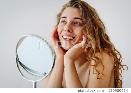 Curly woman touches red inflammations on her face while looking in the mirror in a bright room. Curly woman touches red inflammations on her face while looking in the mirror in a bright room. 120980414