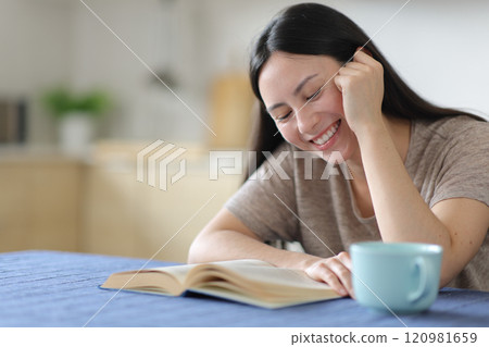 Happy asian woman reading a paper book sitting in the kitchen Happy asian woman reading a paper book sitting in the kitchen 120981659