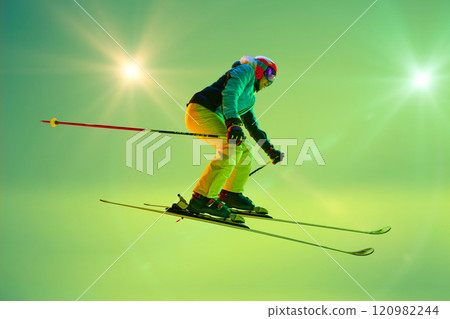Female skier performing jump, dynamic body posture in neon green vibrant background with glowing effect and sun beams Female skier performing jump, dynamic body posture in neon green vibrant background with glowing effect and sun beams 120982244